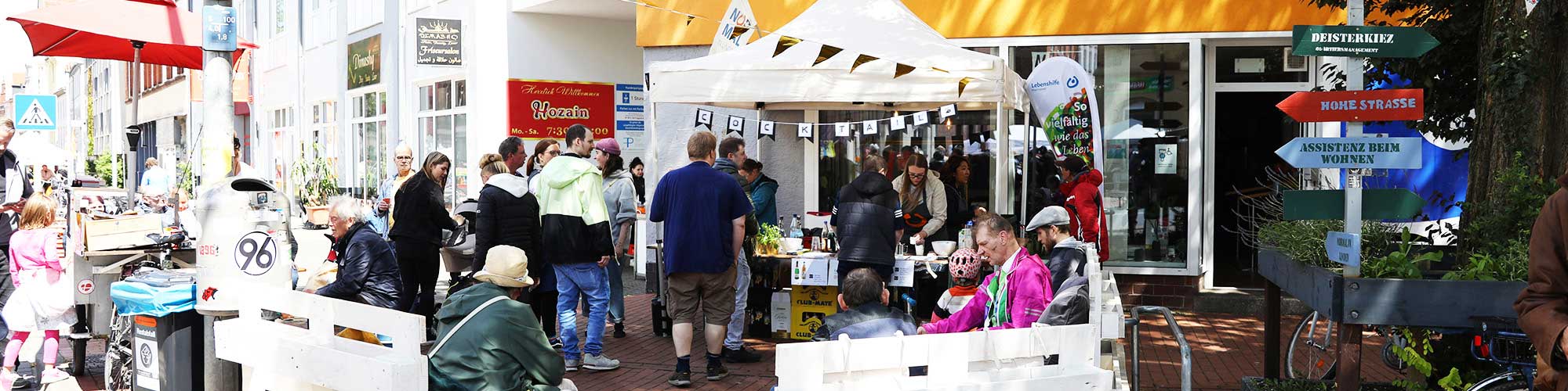 Bildbeschreibung Headerbild: Gezeigt wird der Eingang zum NiL. Vor dem Eingang steht ein Stand, an dem Cocktails gemixt und verkauft werden. Darum herum stehen Bänke, auf denen Leute sitzen.