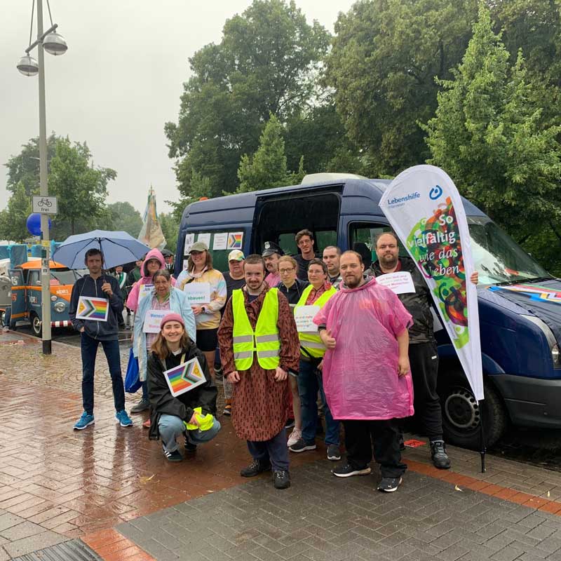 Bildbeschreibung: Vor einem dunklen Transporter stehen einige Personen und lächeln in die Karmera. Ein paar haben Regenumhänge an. Rechts sieht man eine Fahne mit dem Lebenshilfe Schriftzug.