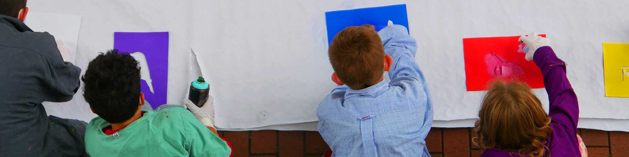 Bildbeschreibung Headerbild: Vier Kinder sitzen vor einer großen weißen Fläche aus Papier und sprühen mit bunten Farben und Schablonen Formen auf das Papier.