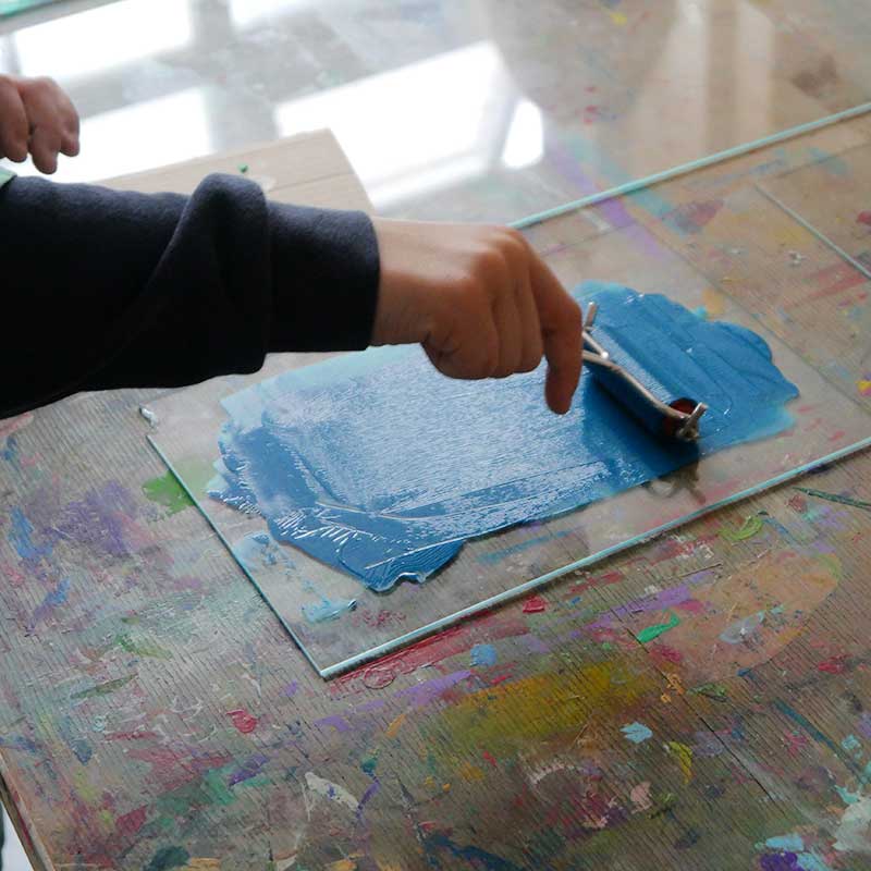 Bildbeschreibung: Gezeigt wird eine Hand, die eine kleine Farb-Rolle hält. Mit der Rolle wird blaue Farbe auf einer Glasscheibe ausgerollt. Bildbeschreibung: Man sieht eine Hand, die eine Rolle hält. Mit der Rolle wird blaue Farbe auf einer Glasscheibe ausgerollt.
