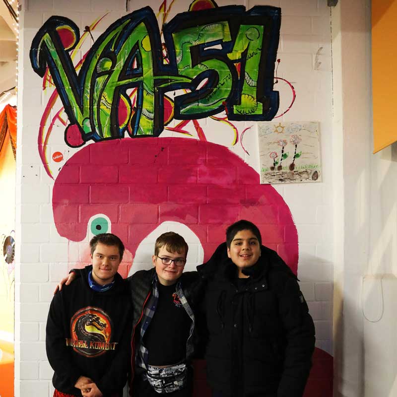 Bildbeschreibung: Drei Jugendliche stehen vor einer Wand mit einem aufgesprayten Logo des ViA 51 . Alle drei lächeln.