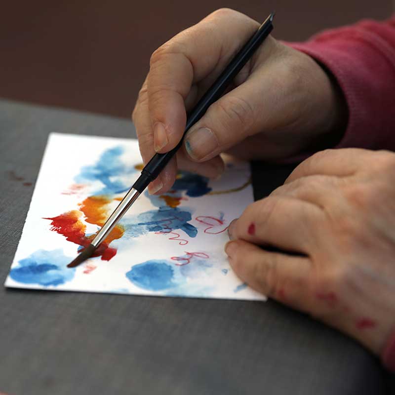 Bildbeschreibung: Eine Hand hält einen Pinsel und malt damit mit Wasserfarben auf Papier. Atelier Charlotte.