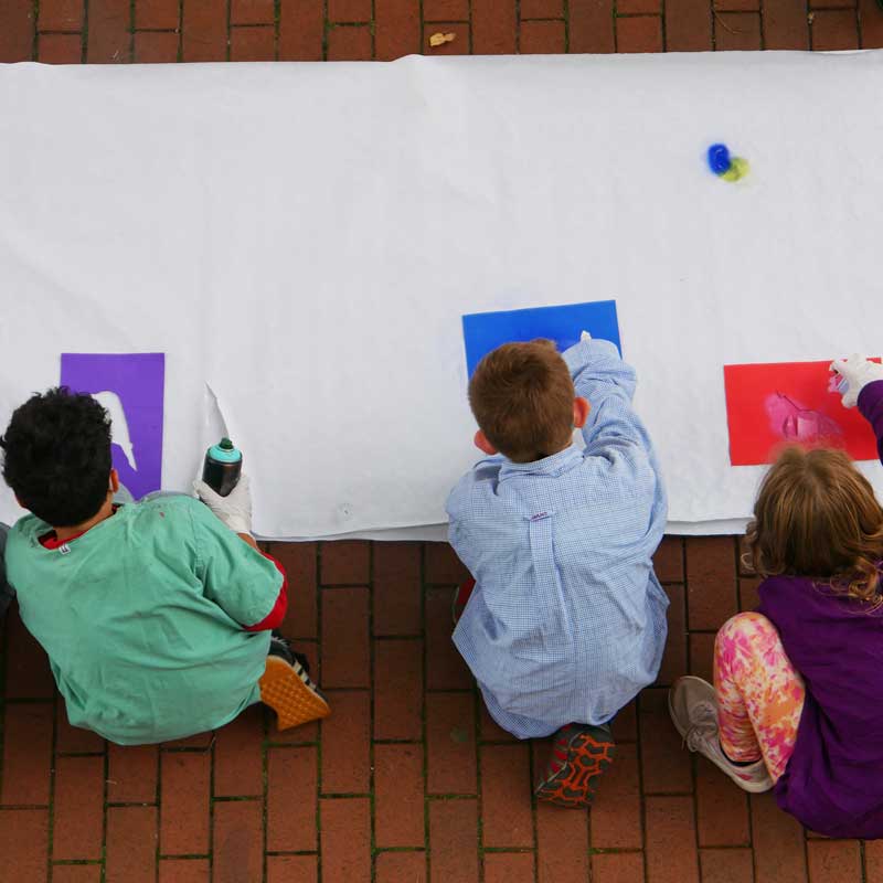 Bildbeschreibung: Drei Kinder sitzen auf dem Boden vor einem riesigen Stück Papier und sprühen mit Sprühfarbe und Schablonen Muster auf das Papier. Finja-Angebote für Kinder und Jugendliche.