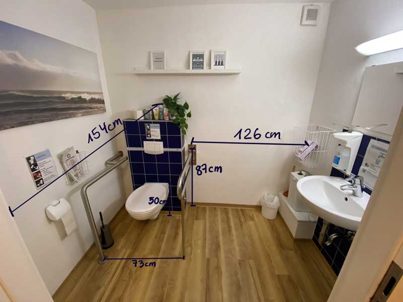 Die große NiL-Toilette mit Maßen Bildbeschreibung: Der große Toilettenraum hat ein Waschbecken und eine Toilette. Die Toilette hat rechts einen Haltegriff und links einen Stützklappgriff.