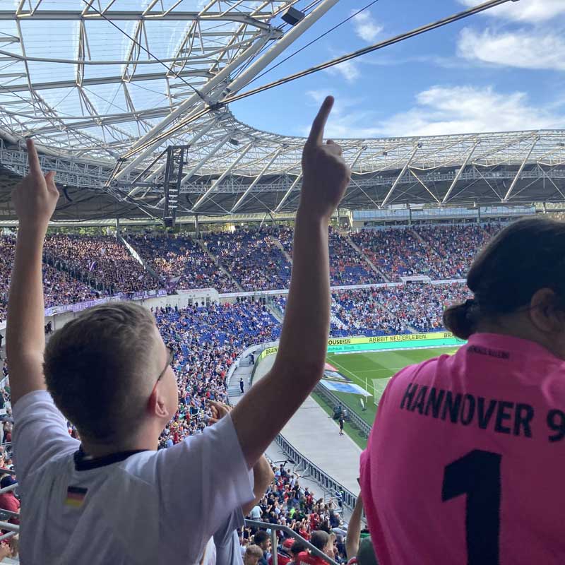 Bildbeschreibung: Das Foto wurde im Stadion von Hannover 96 gemacht. Im Stadion sind viele Menschen. Auf dem Foto sieht man zwei Personen von hinten. Eine Person jubelt. Finja-Angebot: Finja-Mobil