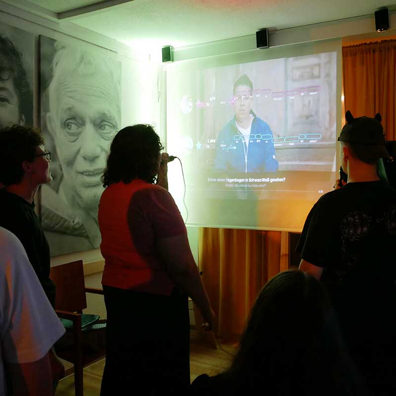Bildbeschreibung: Auf dem Bild sind mehrere Personen zu sehen. Zwei Personen singen gemeinsam Karaoke während die andere zusehen und zuhören. Im Hintergrund ist eine Leinwand, auf der der Text und die Tonhöhe zu sehen ist. Finja-Angebot: Junges Event-Team