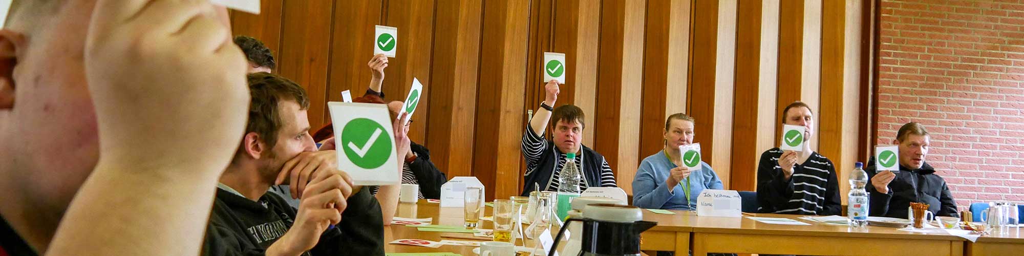 Bildbeschreibung Headerbild: Viele Leute sitzen in einem Raum und stimmen ab. Sie halten kleine Karten hoch, auf denen entweder ein grüner Kreis mit einem weißen Haken oder ein roter Kreis mit einem weißen X ist. Der grüne Kreis bedeutet "Ich stimme zu", der rote Kreis bedeutet "Ich stimme nicht zu".